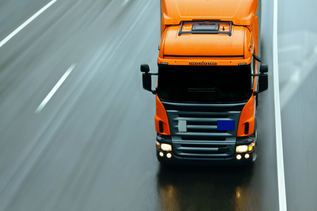 Overhead view of an orange lorry driving along a multi‑lane road, captured from above, with headlights on and road markings visible beneath.