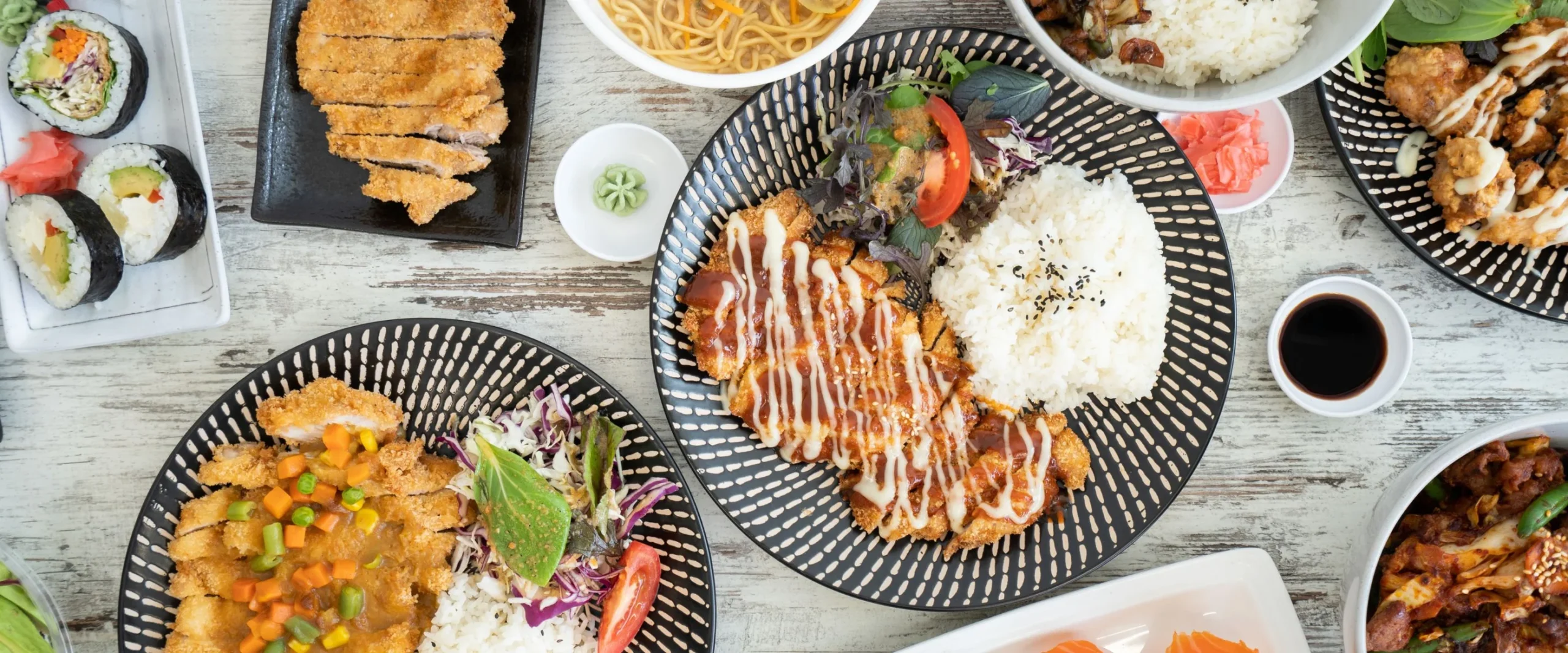 A colourful spread of fresh, beautifully plated Asian-inspired dishes, including crispy katsu, steamed rice, sushi rolls, noodles, and vibrant salads, arranged on patterned plates for an appetising, restaurant-style presentation.