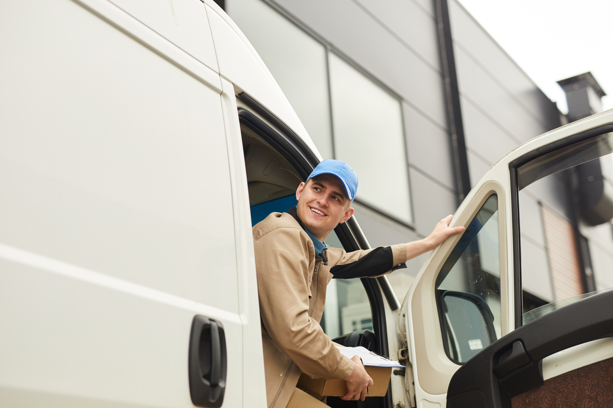 Young smiling courier holding package while sitting in the van he delivering parcels