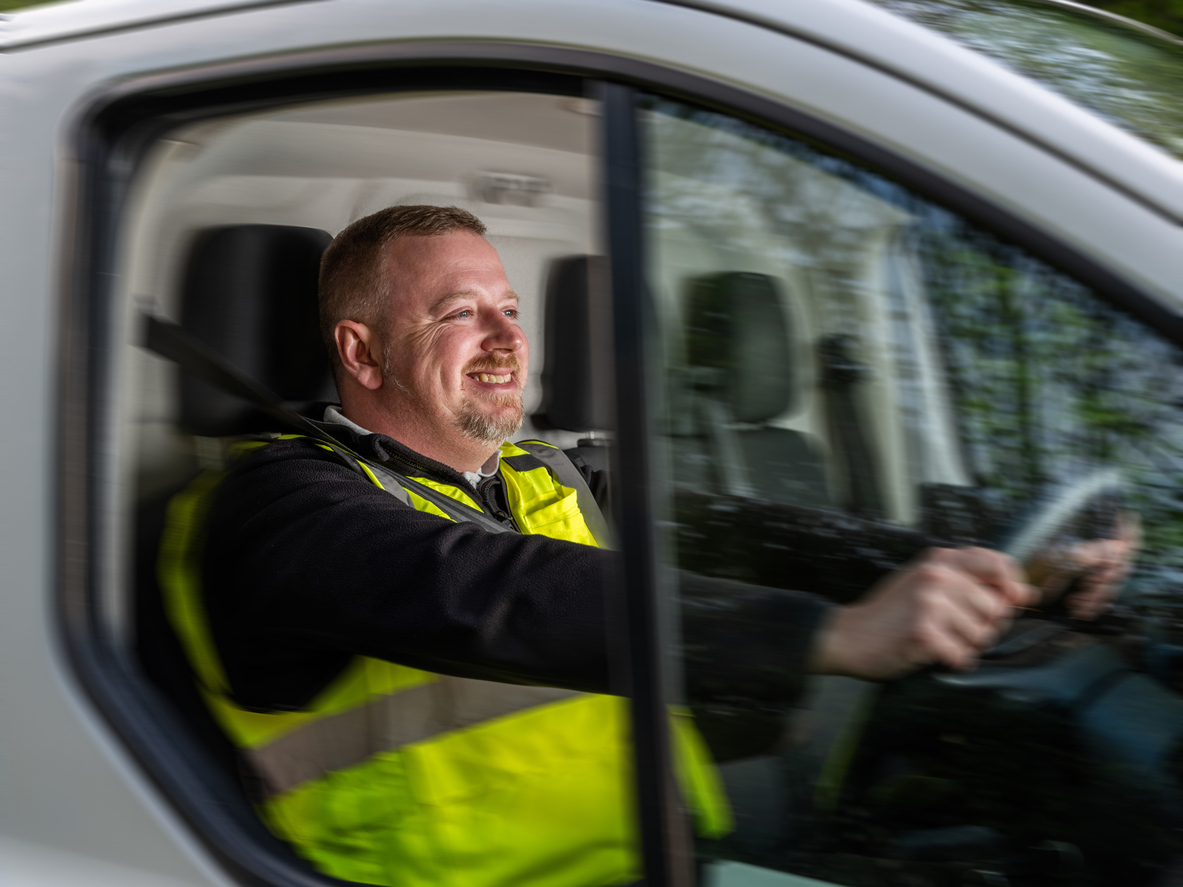 A cheerful van driver, dressed in a high-visibility vest, is captured steering a white van while in motion, smiling brightly during daylight. The blurred greenery outside the window highlights the vehicle’s movement, with natural sunlight illuminating the passenger cabin. This image, showcasing the driver’s positive expression, is ideal for illustrating transportation, workplace positivity, or the daily life of a delivery person in the transport occupation.