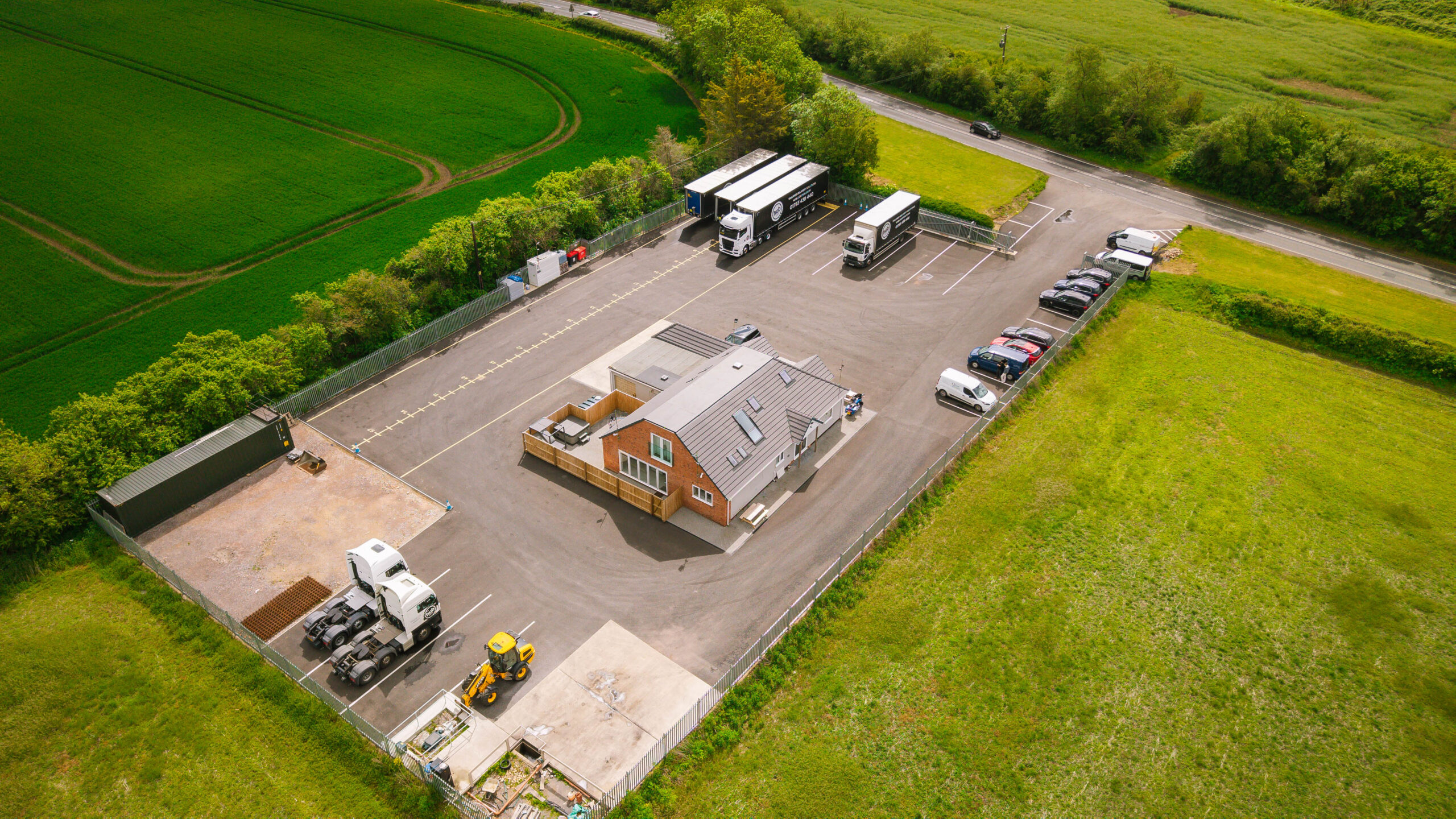 Aerial view of a small industrial yard: brick office building, loading bays, several parked semi-trailers, and surrounding green fields and trees.
