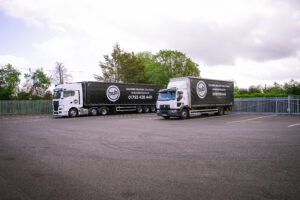 Two large black delivery trailers with hub Logistics branding parked in a lot, white cabs attached, behind a metal fence and trees under a cloudy sky