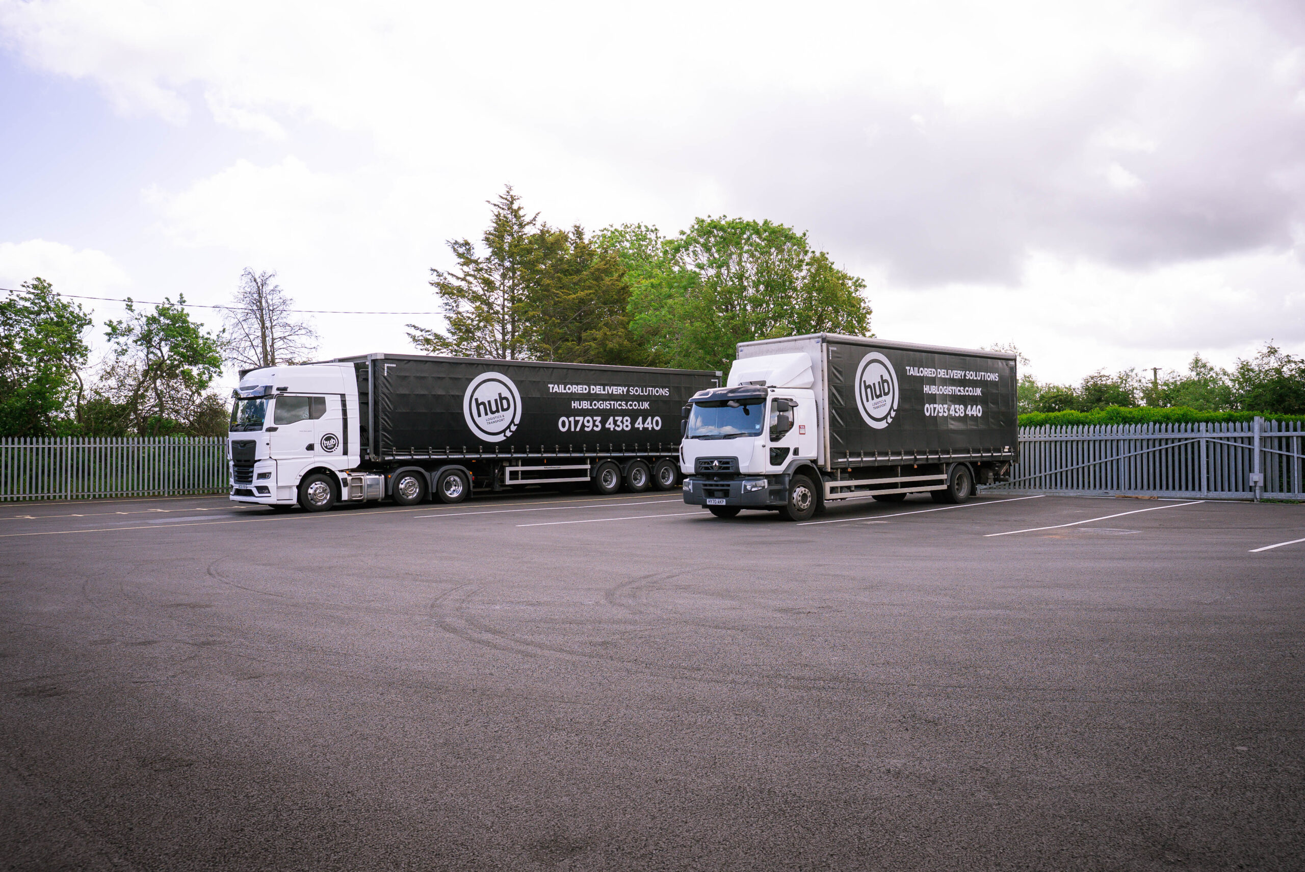 Two large black delivery trailers with hub Logistics branding parked in a lot, white cabs attached, behind a metal fence and trees under a cloudy sky