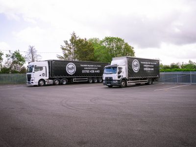 Two large black delivery trailers with hub Logistics branding parked in a lot, white cabs attached, behind a metal fence and trees under a cloudy sky