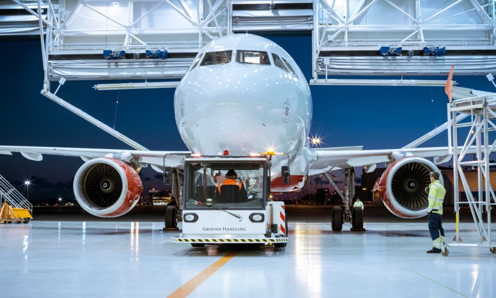 Aircraft Maintenance Hangar Where New Airplane is Toed by a Pushback Tractor/ Tug onto Landing Strip. Crew of Mechanics, Engineers and Drivers Works Busily.
